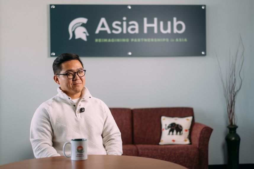 Young male professor smiles and sits calmly in front of Asia Hub mug
